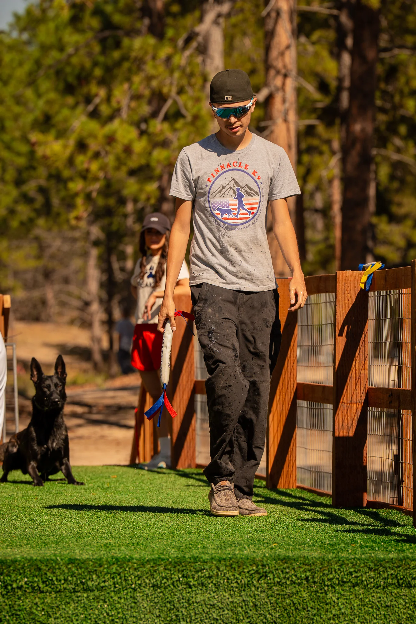 Handler carrying a bumper with a seated black dog behind.