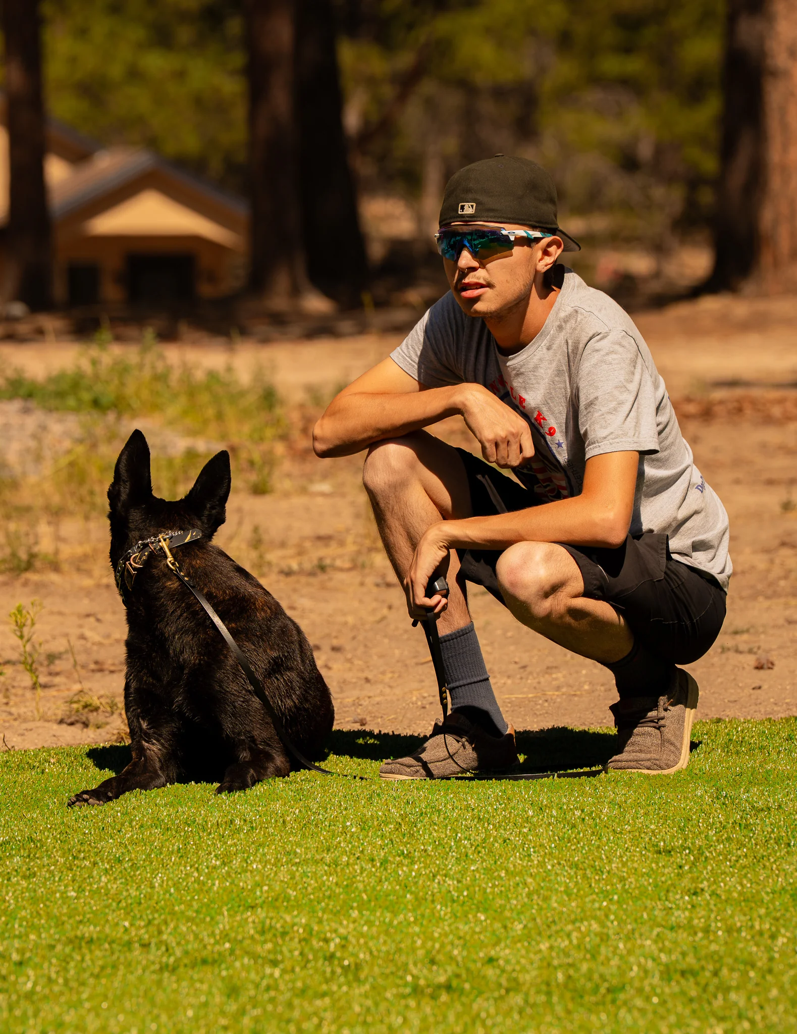 Handler crouching beside a seated black dog on turf.
